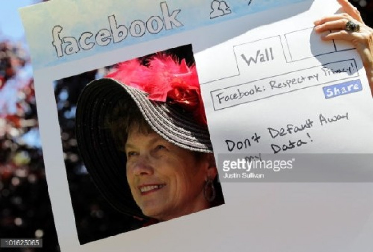 Photo of a Woman in a large hat with feathers, holding a protest sign asking Facebook not to give away users privacy by default