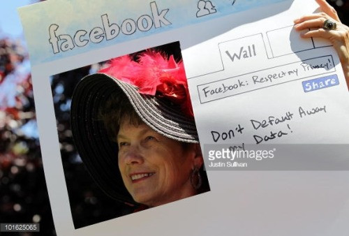 Photo of a Woman in a large hat with feathers, holding a protest sign asking Facebook not to give away users privacy by default