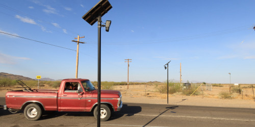 A red truck carrying a lawnmower drives past two Flock Safety automated license plate readers. 